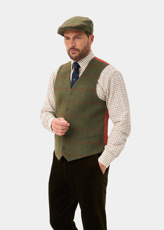 Man in Combrook tweed waistcoat (reed), green flat cap, check shirt, tie & dark trousers stands against plain background.