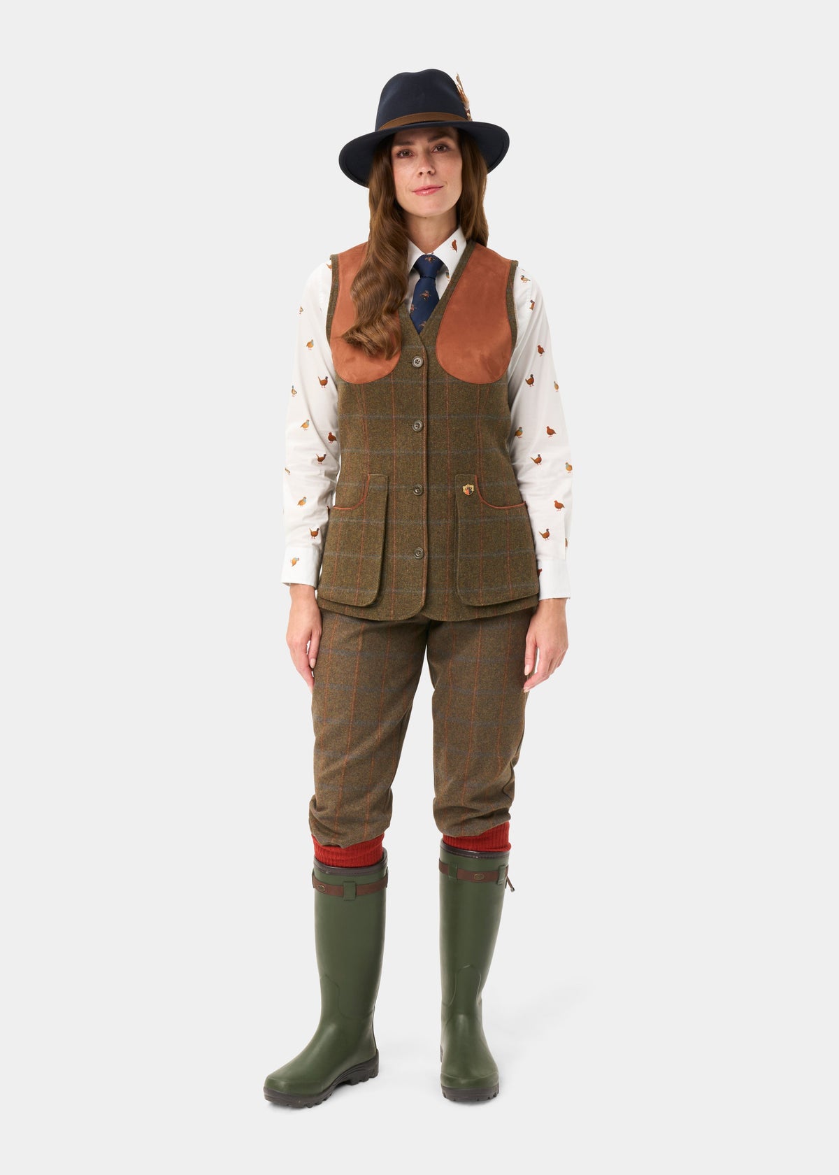 Woman models Combrook Ladies Tweed Shooting Waistcoat in Willow with shirt, tie, hat, green boots, and brown pants.