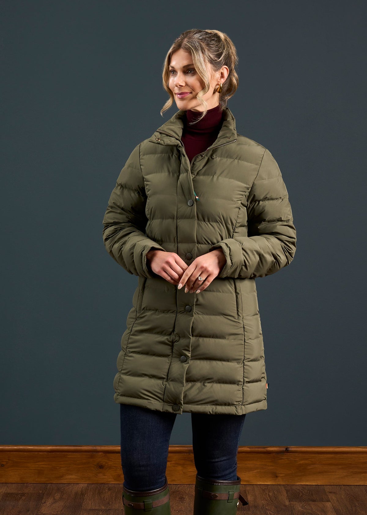 Woman in a Calsall Ladies Coat in Olive - Regular Fit with dark jeans and boots stands before a plain dark background.