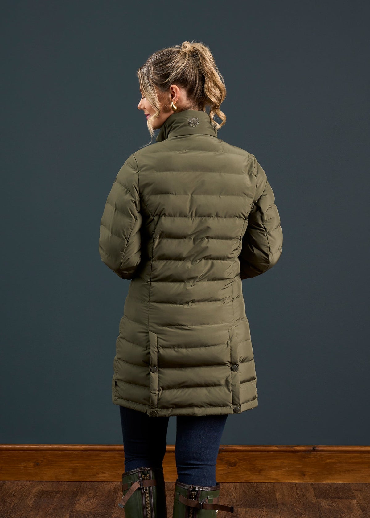 Woman with blonde ponytail wears Calsall Ladies Coat in Olive, Regular Fit, water repellent, facing away from camera.