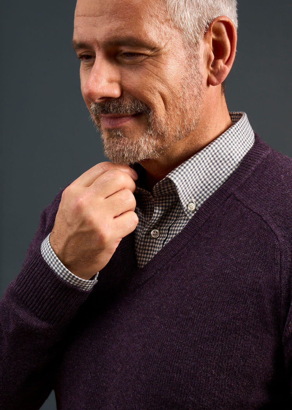Smiling man in Kilsyth Lambswool Vee Neck Jumper in Elderberry adjusts his collar against a dark background.