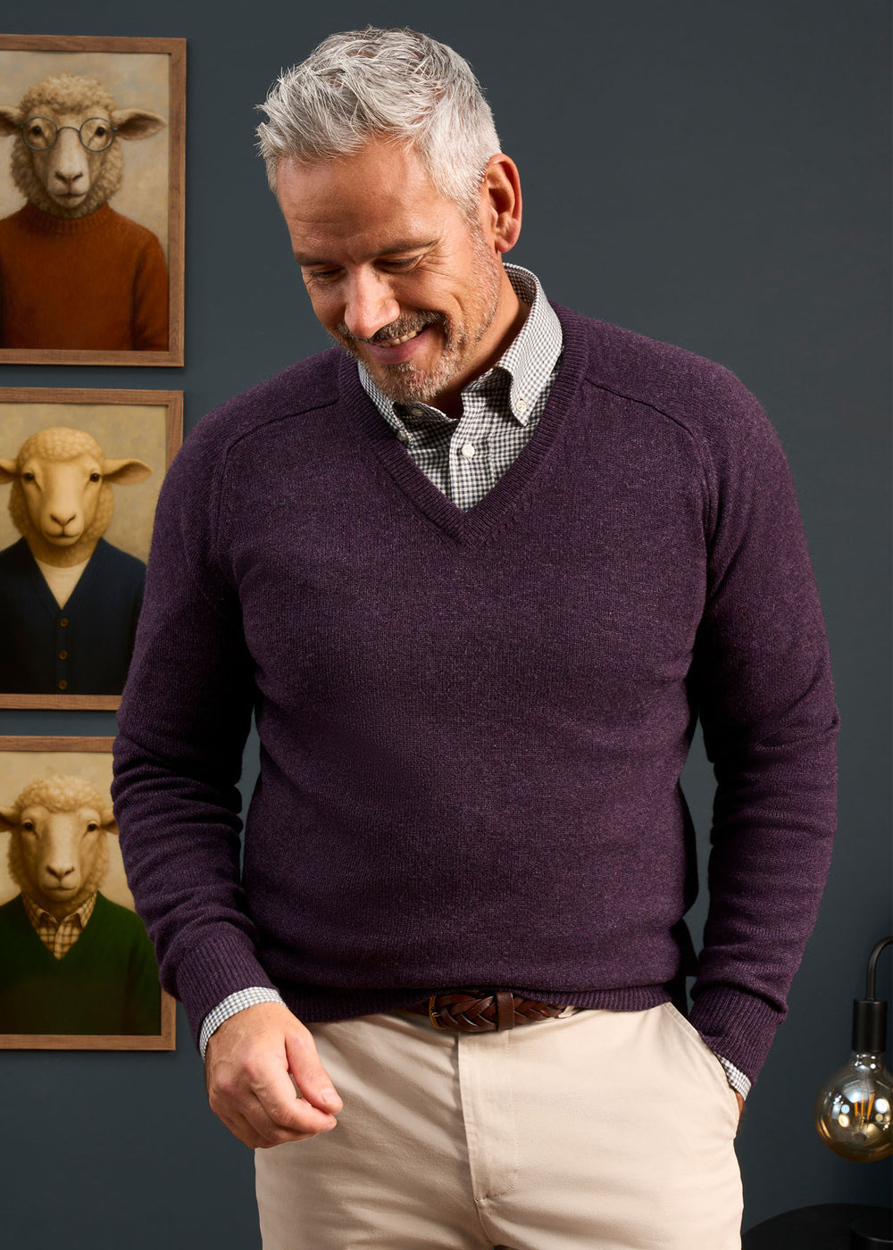 Smiling man wears Kilsyth Lambswool Vee Neck Jumper in Elderberry, standing before sheep portraits on a dark wall.