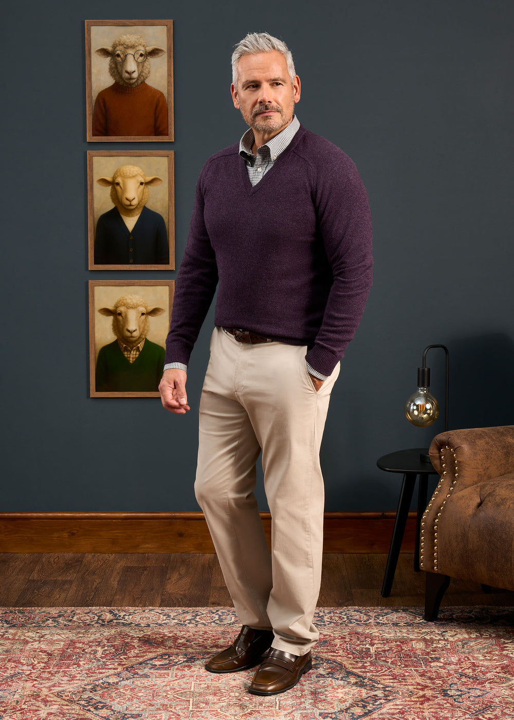 A man in a Kilsyth Lambswool Vee Neck Jumper in Elderberry stands by sheep portraits and a brown chair.