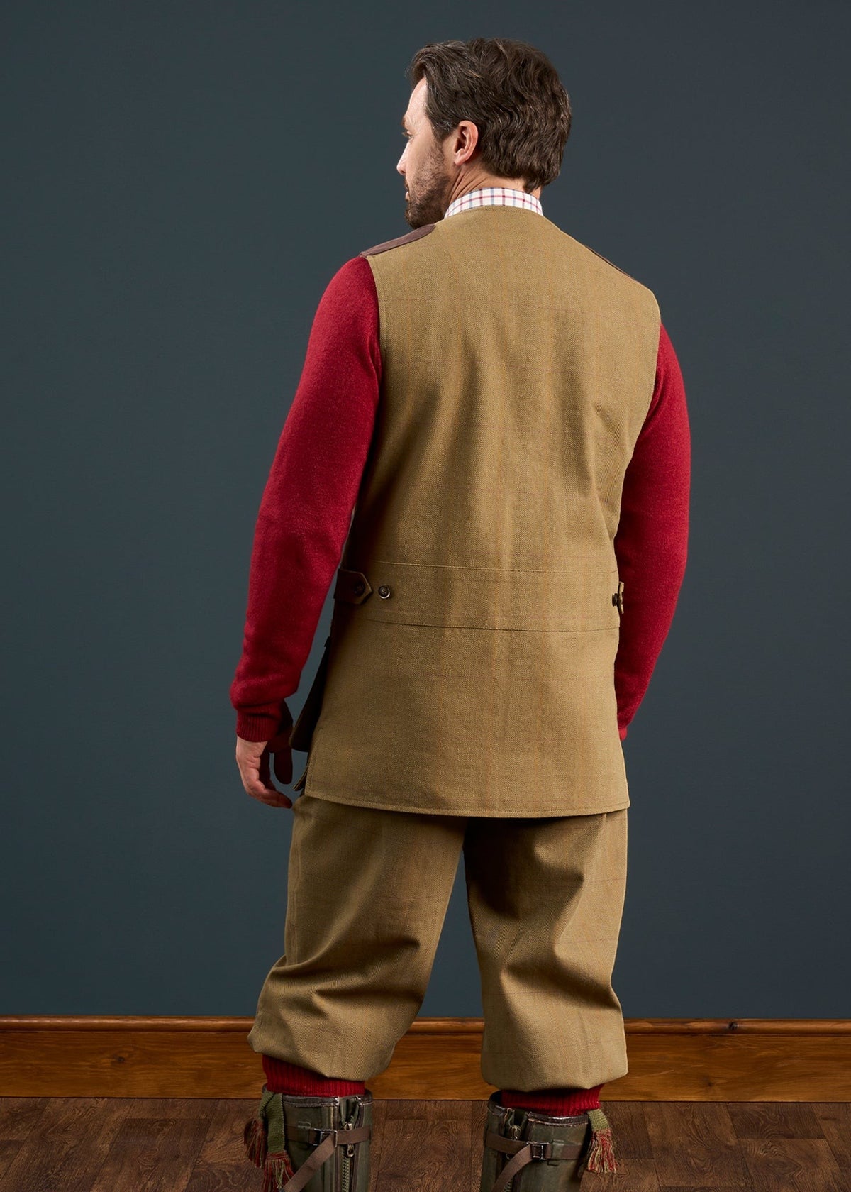 Man facing away in Axford Men’s Waterproof Lightweight Shooting Waistcoat, tan pants, red sweater, boots, on wood floor.