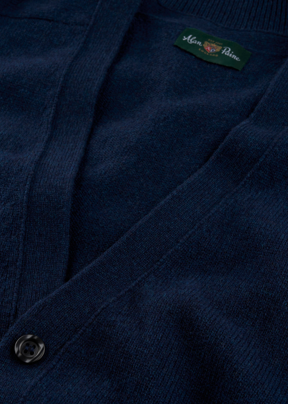 Close-up of the Cornwall Lambswool V-Neck Cardigan in Indigo with black button and green Mina Paine label.