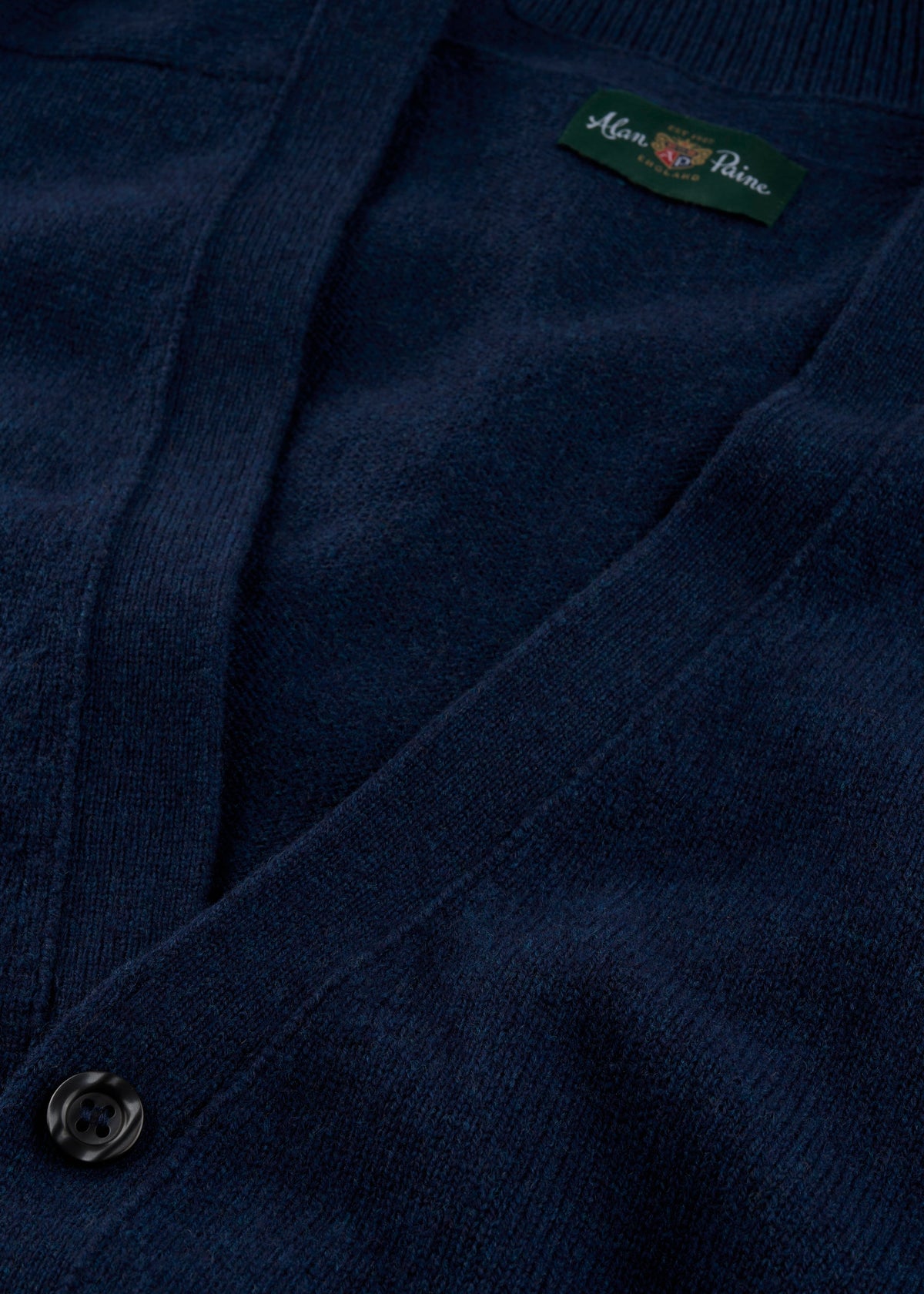 Close-up of the Cornwall Lambswool V-Neck Cardigan in Indigo with black button and green Mina Paine label.