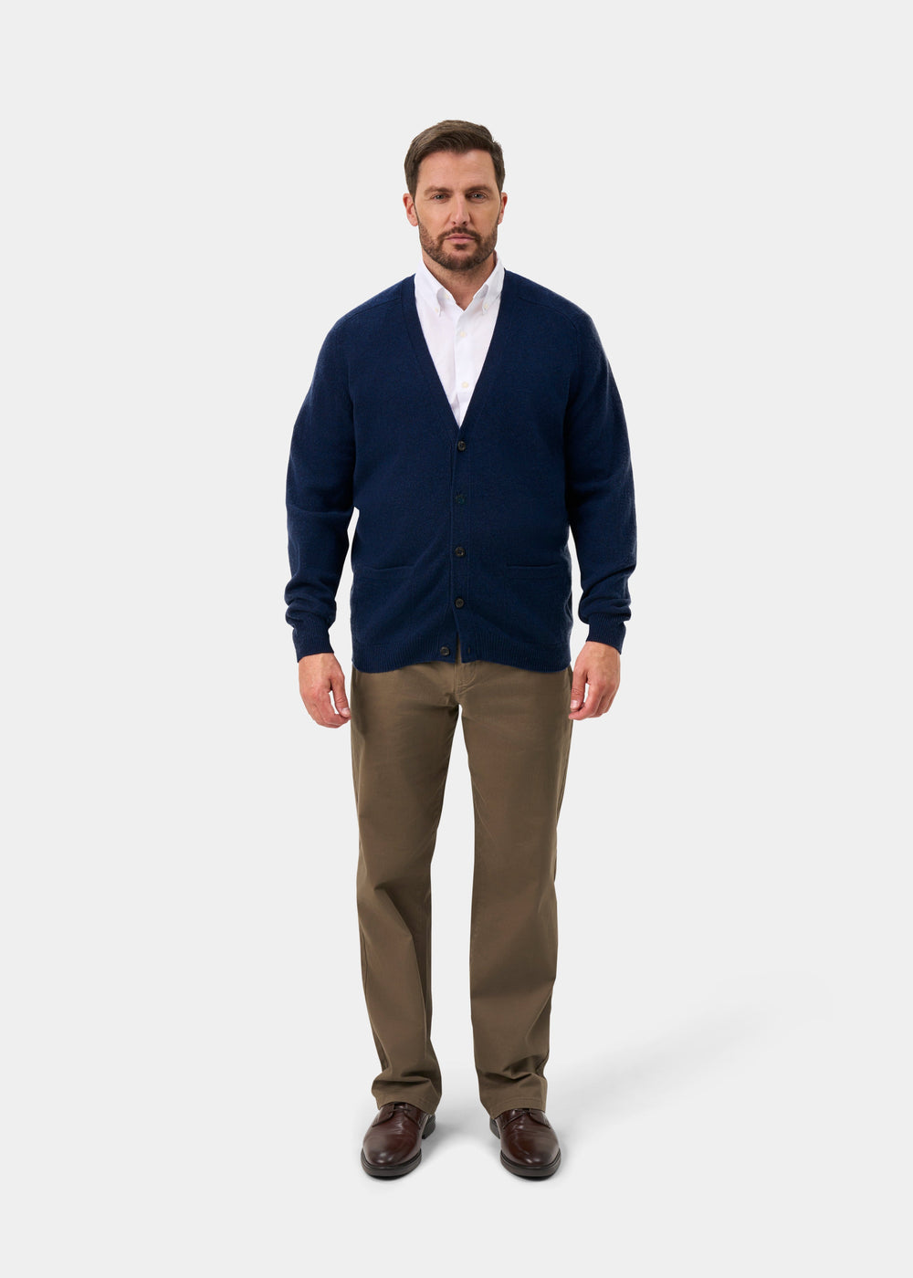 Man in Cornwall Lambswool V-Neck Cardigan in Indigo, white shirt, khaki pants, brown shoes, plain background.