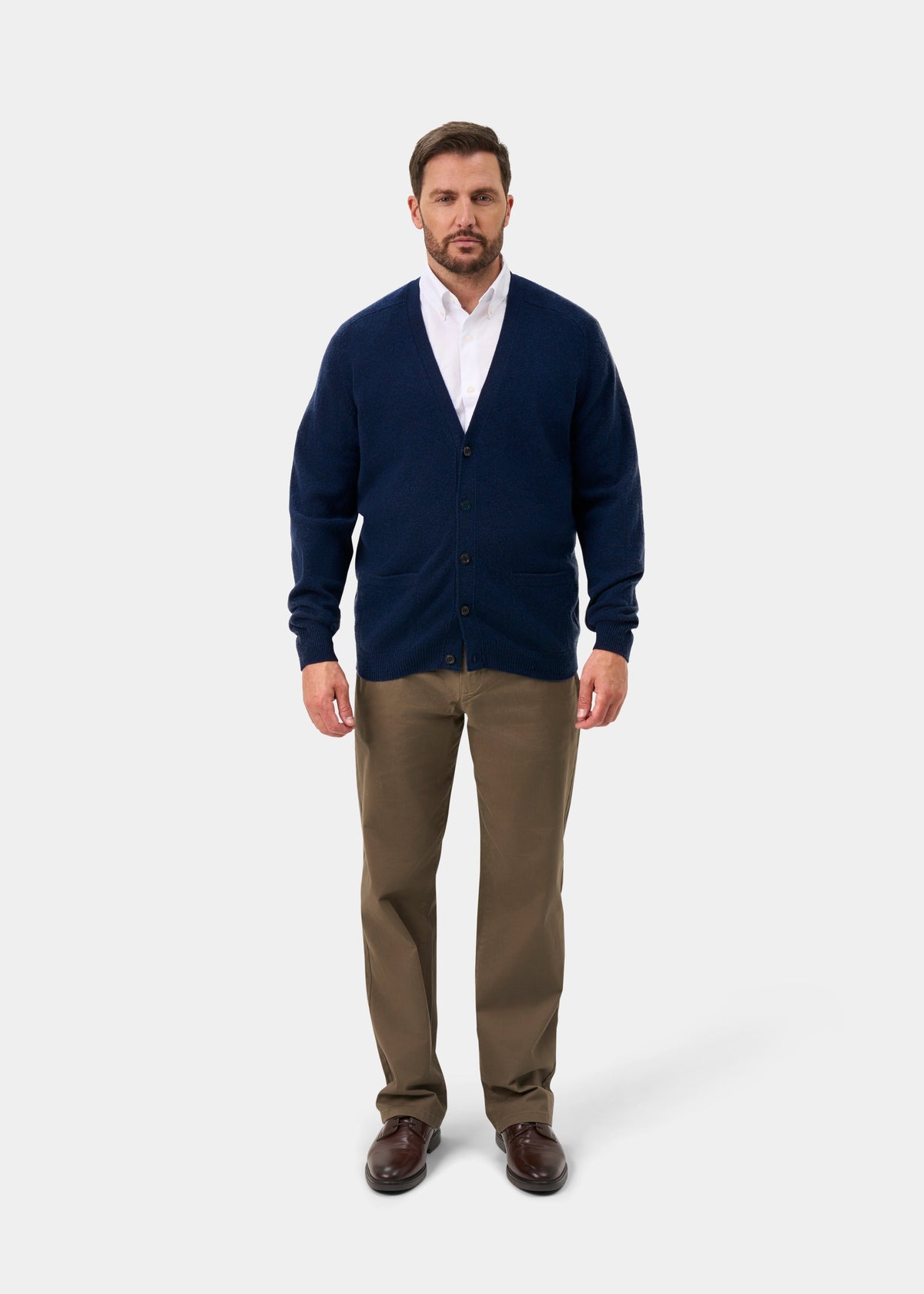 Man in Cornwall Lambswool V-Neck Cardigan in Indigo, white shirt, khaki pants, brown shoes, plain background.