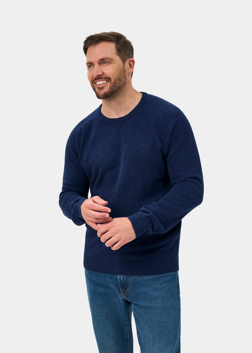 Smiling man wears the Lenzie Lambswool Crew Neck Jumper in Rhapsody with blue jeans, standing by a plain gray backdrop.