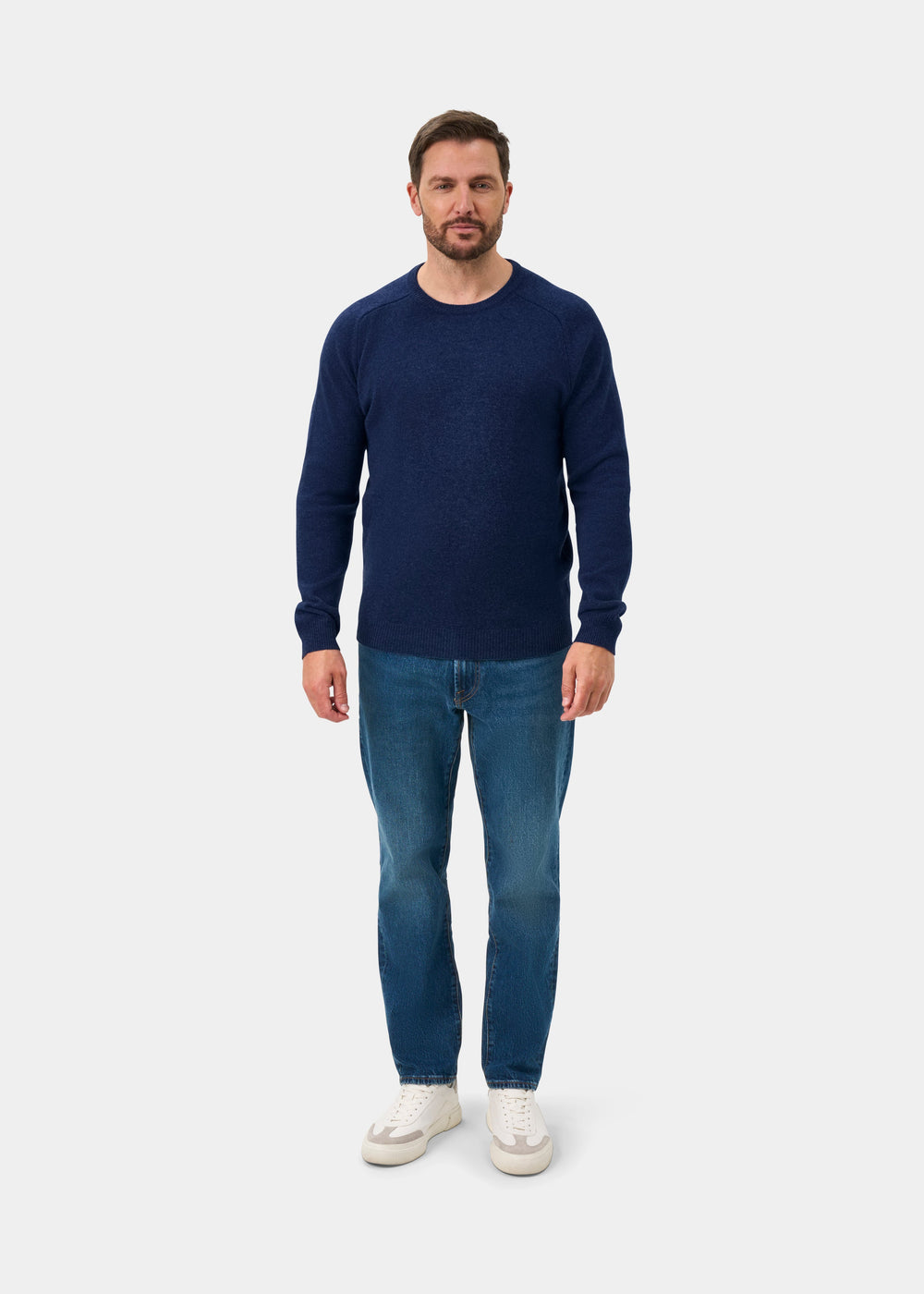 Man in Lenzie Lambswool Crew Neck Jumper in Rhapsody, blue jeans, and white sneakers stands on white background.