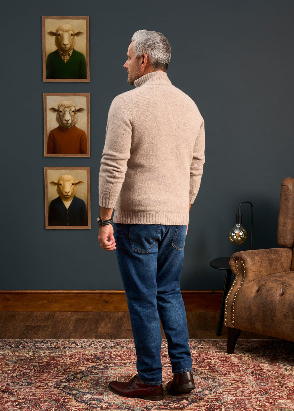 A man in a Landford Lambswool Buttoned Mock Neck Jumper gazes at sheep portraits, standing on a patterned rug.
