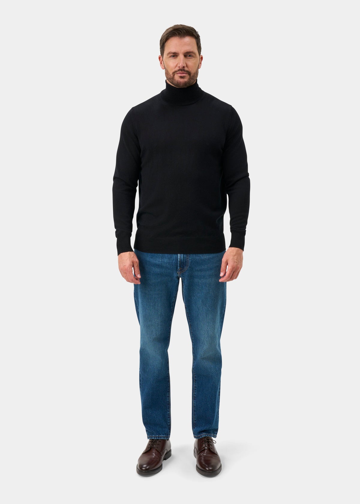 Man wears the Linton Merino Wool Roll Neck Jumper in black, blue jeans, and brown shoes against a plain background.