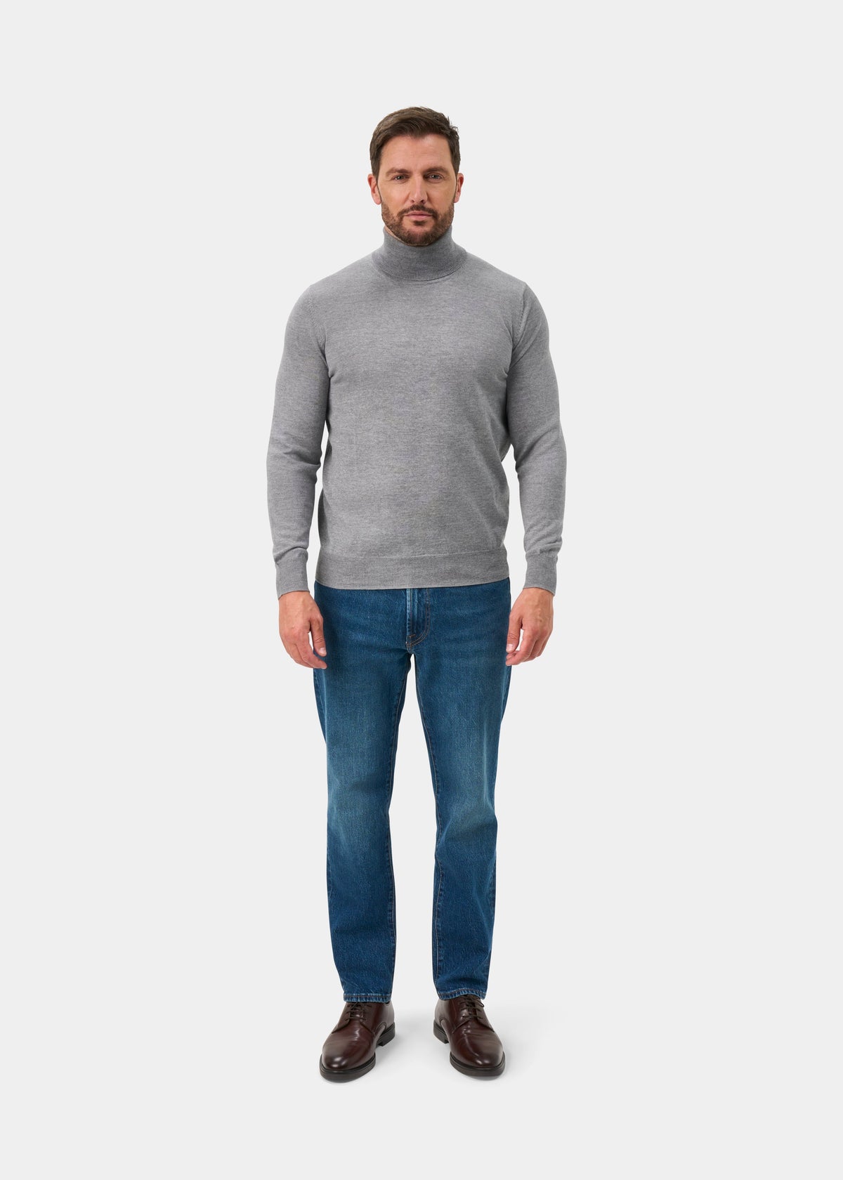 Man wears Linton Merino Wool Roll Neck Jumper in light grey mix, blue jeans, and brown shoes against a white background.