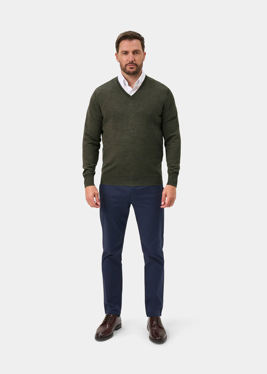 A man modeling a dark olive green v-neck sweater over a white collared shirt, paired with navy blue trousers and brown leather shoes, against a light gray studio background.