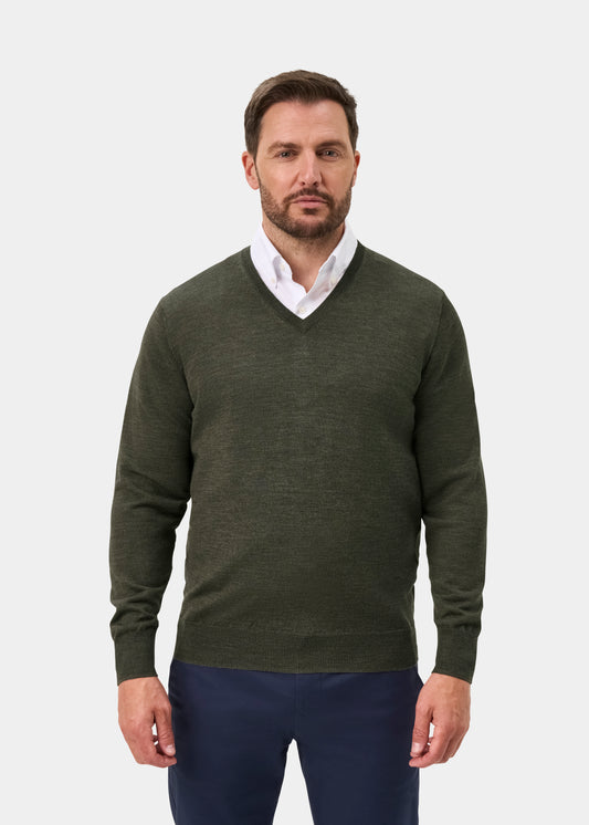 A man wearing an olive green v-neck sweater over a white collared shirt and navy blue trousers, standing against a light gray studio background.