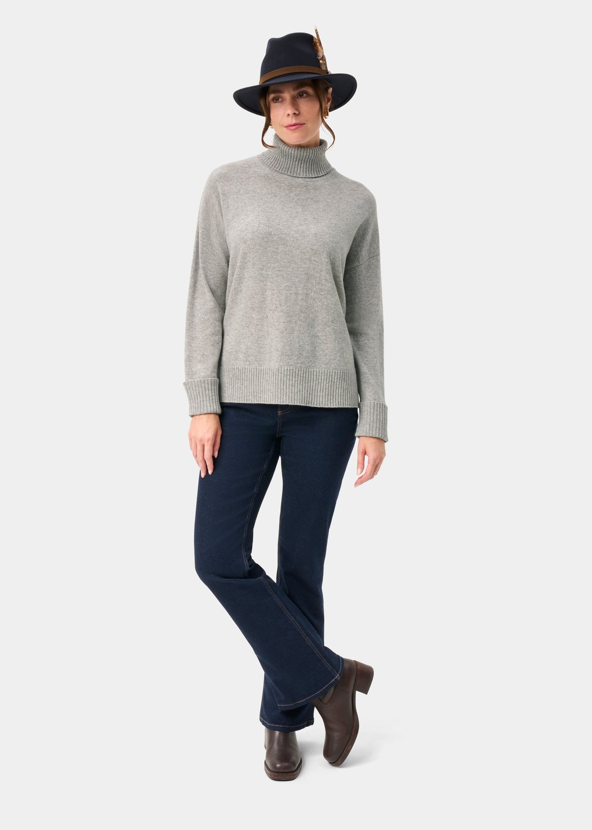 Woman in Alexa Wool & Cashmere Roll Neck Jumper in Husky, dark jeans, brown boots, and black feather hat.