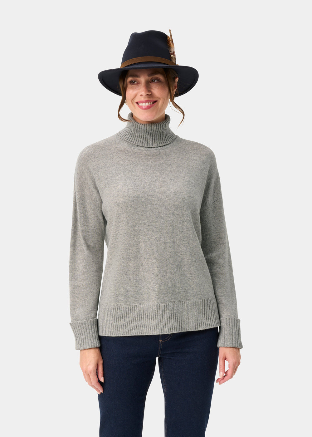 Woman in the Alexa Wool & Cashmere Roll Neck Jumper in Husky, smiling in a dark feathered hat against a white background.