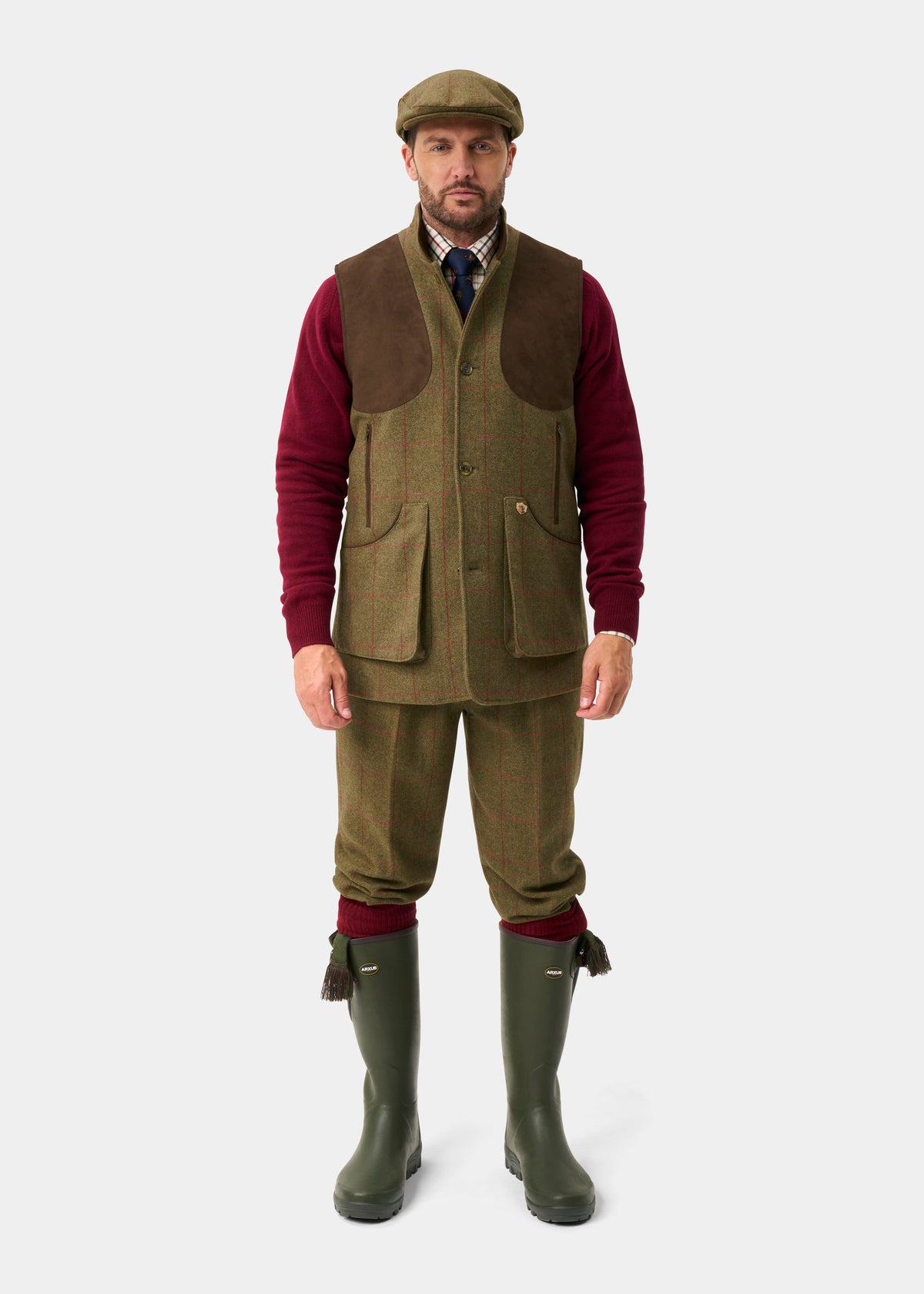 Man in Combrook Men’s Tweed Water Repellent Shooting Gilet in Sage, maroon sweater, green boots, brown cap, white backdrop.