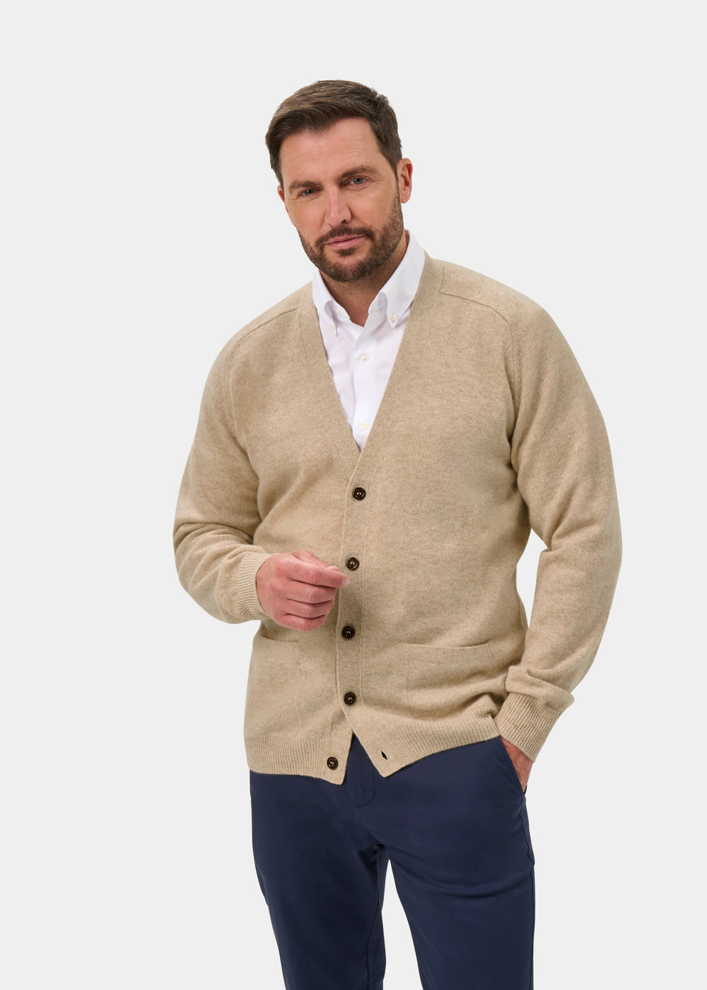 Man wears Cornwall Lambswool V-Neck Cardigan in Linen, white shirt, navy pants; stands by a plain white background.