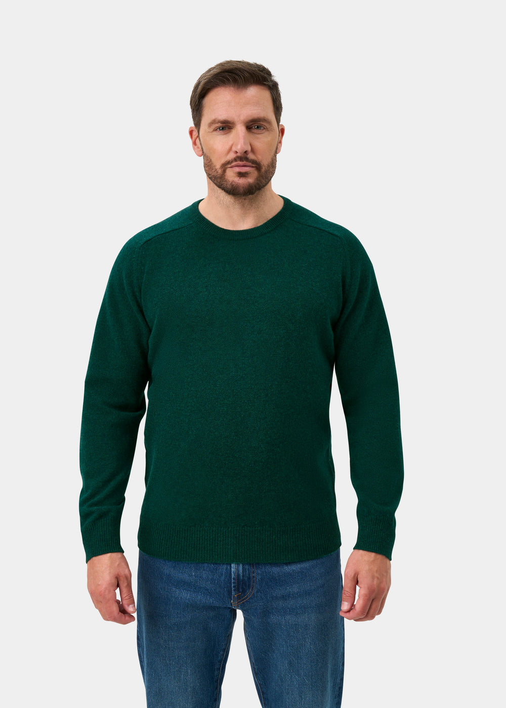 Man in Dorset Lambswool Crew Neck Jumper in Mallard and blue jeans stands against a plain white background.