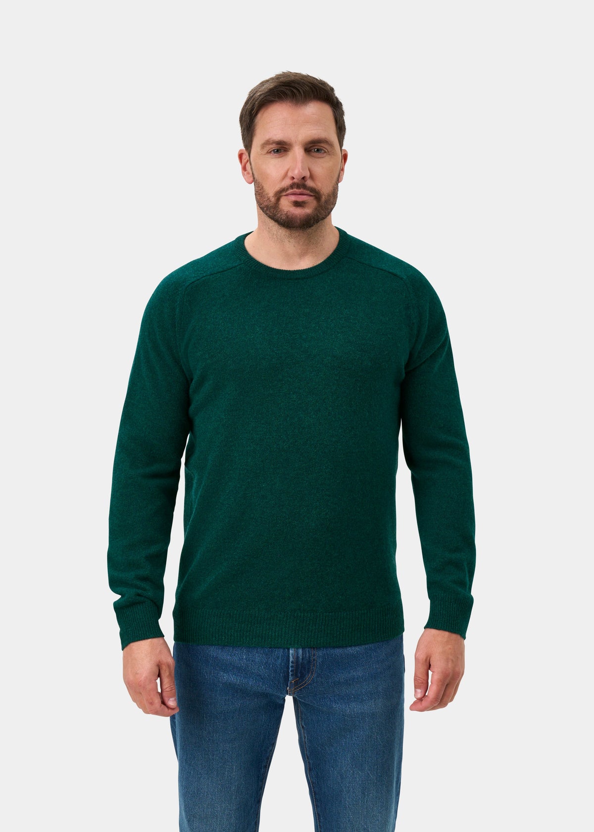 Man wears the Lenzie Lambswool Crew Neck Jumper in Mallard with blue jeans, standing against a plain white background.