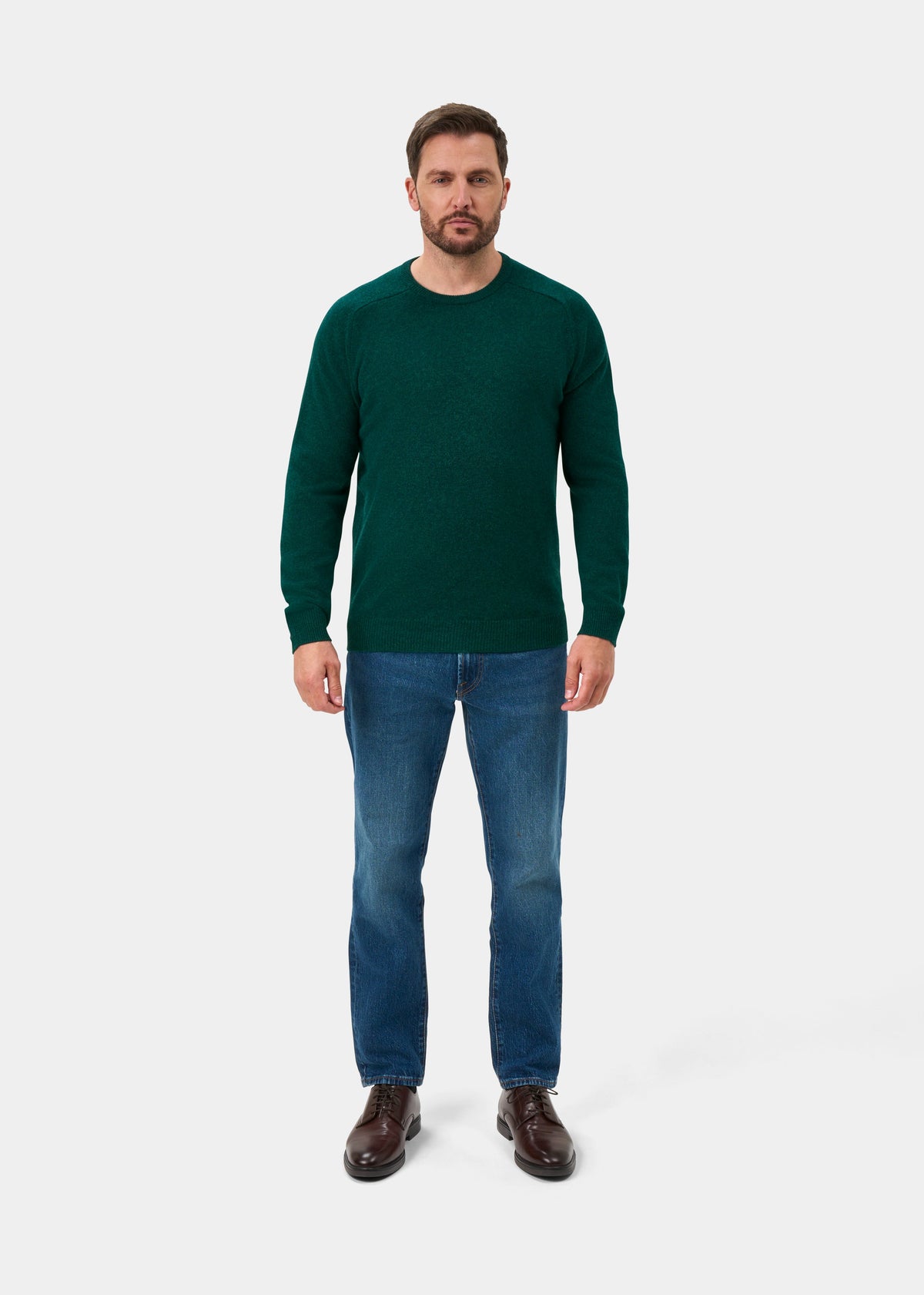 Man facing forward in Lenzie Lambswool Crew Neck Jumper in Mallard, blue jeans, and brown shoes on white background.
