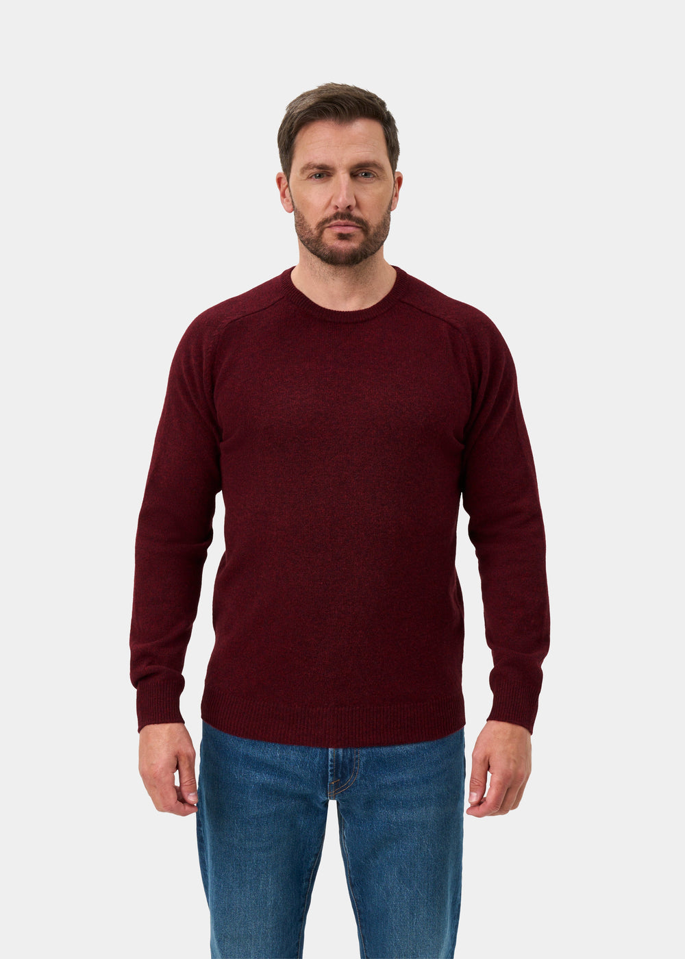 Man wears Lenzie Lambswool Crew Neck Jumper in Red Velvet, regular fit, with blue jeans against a plain white background.