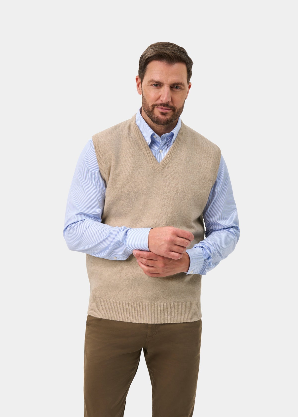 A man wearing a beige knit V-neck sweater vest over a light blue dress shirt, paired with brown trousers, photographed against a light gray studio background.