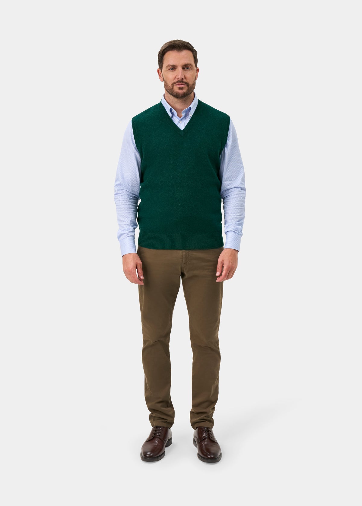 A man modeling a dark green V-neck knit sweater vest over a light blue long-sleeve dress shirt, paired with brown trousers and dark brown leather dress shoes, against a light gray studio background.