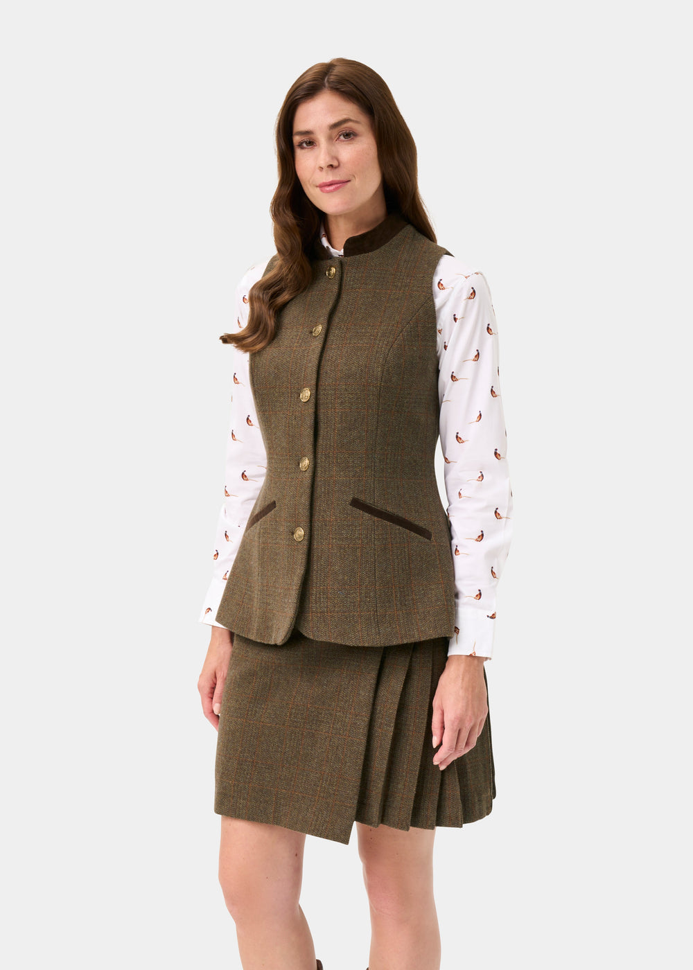 A woman modeling a brown tweed vest and matching pleated skirt over a white patterned long-sleeve blouse, photographed against a light gray studio background.